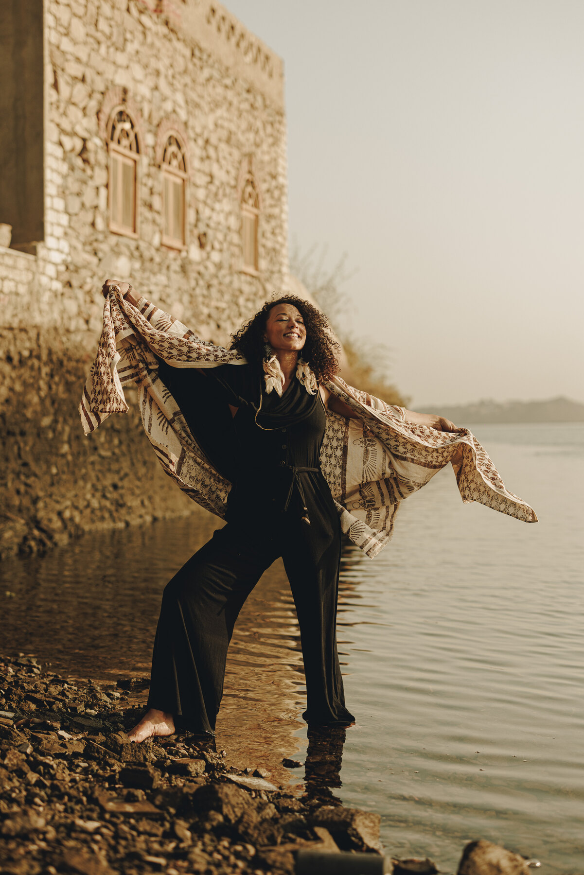 Zai’Ra Paige standing at the water’s edge with her arms open and a flowing shawl